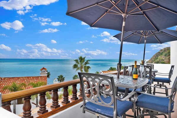 A balcony with a table and chairs, overlooking a coastal view with the sea and palm trees.