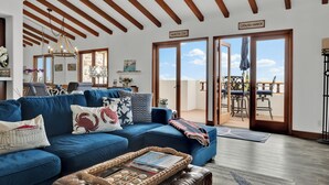 A spacious living room with a blue sofa, wooden ceiling beams, and a view of the balcony.