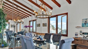 A dining area with a glass table, grey upholstered chairs, a chandelier, and large windows with a view of the sea.