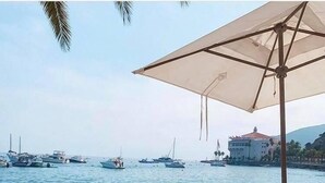 A beach with wooden lounge chairs and a large umbrella, overlooking a calm sea with boats and a building in the background.
