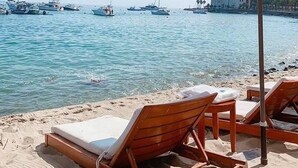 Ein Strand mit Holzliegen und einem großen Sonnenschirm, mit Blick auf ruhiges Wasser, wo Boote und ein Gebäude im Hintergrund zu sehen sind.