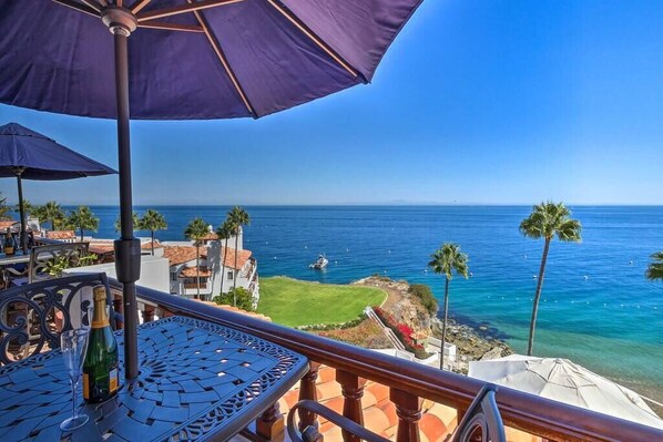 A balcony with a view of the ocean, a table with a bottle, and a purple umbrella. -  Oceanfront Hamilton Cove Penthouse suite with stunning panoramic views! (Avalon)
