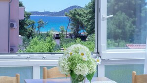 Une table avec un vase de fleurs, une fenêtre donnant sur la mer et les montagnes, et une chaise.