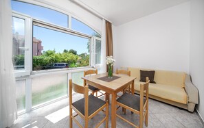 A modern living room with a wooden table and chairs, a beige sofa, and a large window offering a view of the street and greenery.