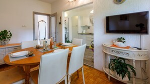 Un comedor con una mesa de madera, sillas blancas, un televisor montado en la pared y una planta en maceta sobre una consola.