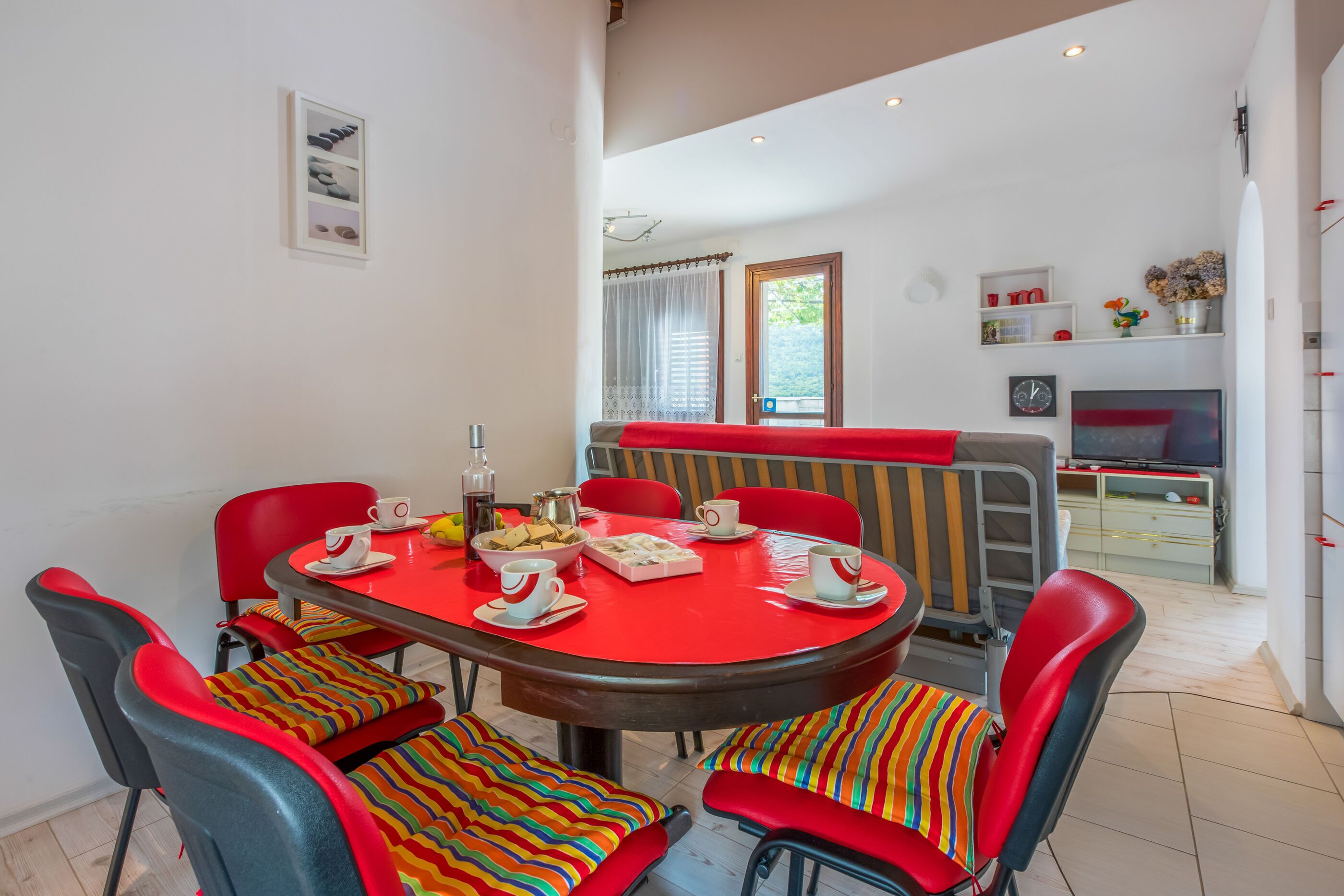 A dining area with a round table, red chairs, and a red tablecloth. There are chairs with colorful striped cushions. A television is mounted on the wall in the background.