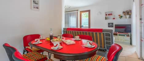 A dining area with a round table, red chairs, and a red tablecloth. There are chairs with colorful striped cushions. A television is mounted on the wall in the background.