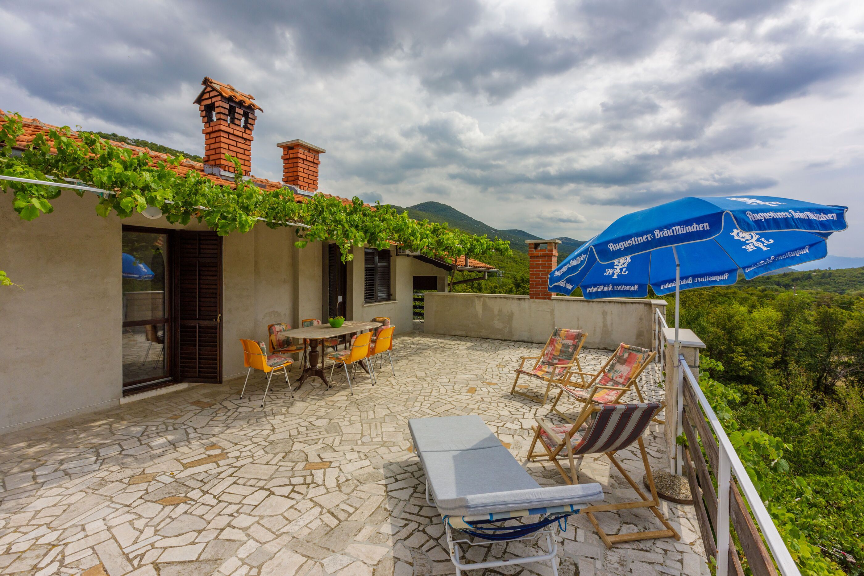 An outdoor patio with stone flooring, a table with chairs, a lounge chair, and a blue umbrella.