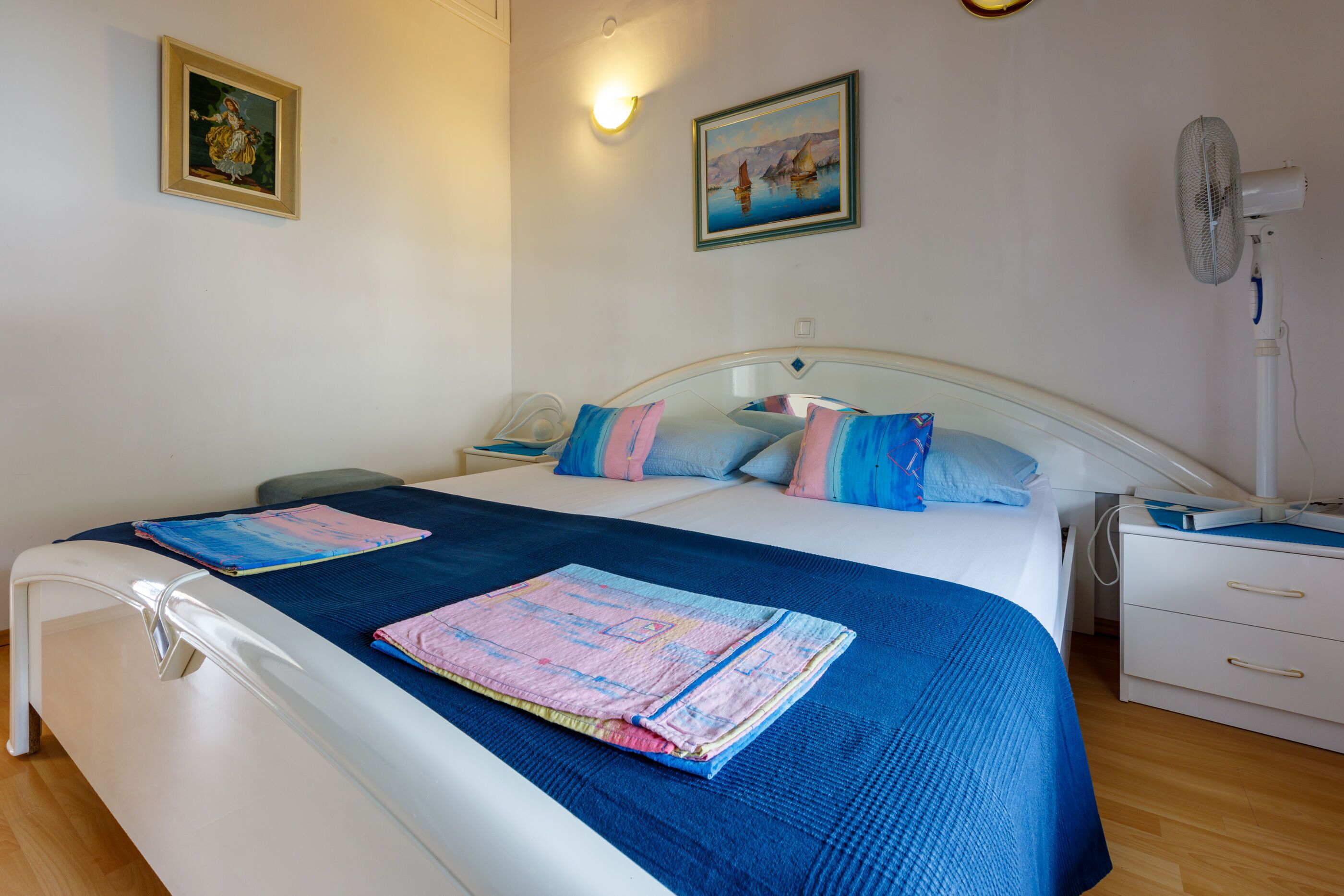 A neatly made bed with a blue and pink quilt, a white bedside table, and a nightstand with a lamp.