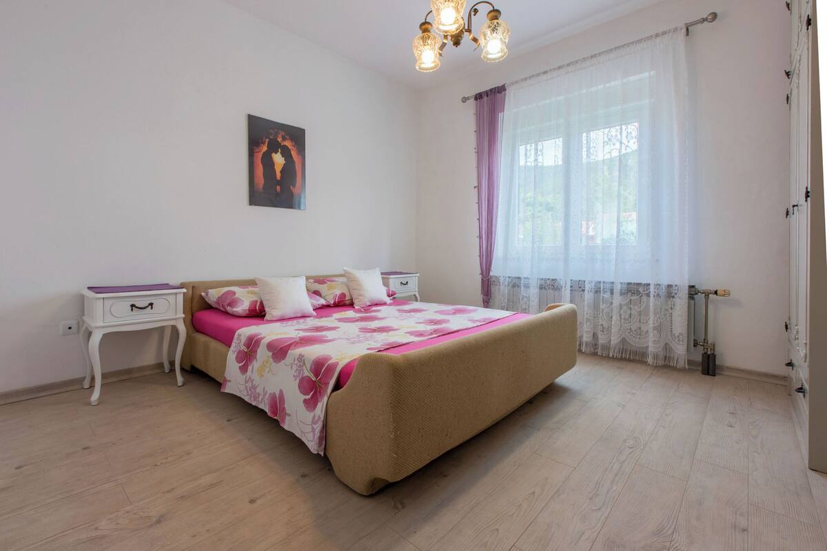 Ein Schlafzimmer mit Bett, Nachttischen, einem Fenster mit Vorhängen und einer Wandleuchte.