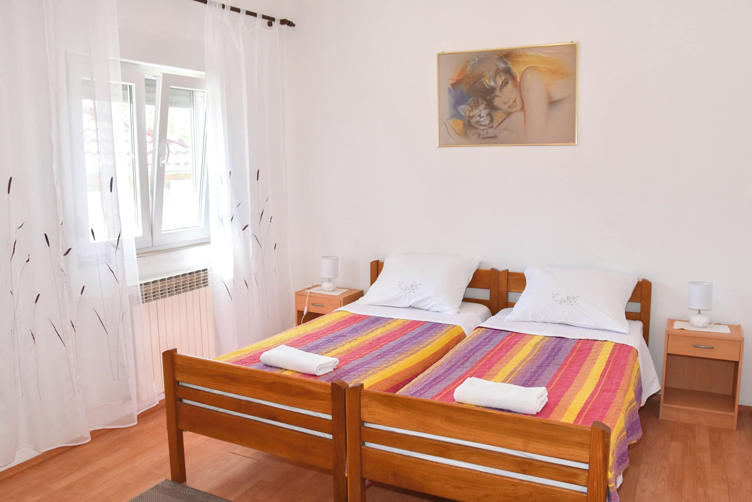 Una cama doble con una colcha a rayas, dos mesitas de noche con lámparas, una ventana con cortinas traslúcidas y un cuadro en la pared.