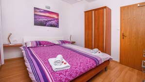 A neatly made bed with a purple and white patterned bedspread, a wooden wardrobe, and a wall-mounted painting of a lavender field. - Apartment Seafront Apartments (Kalebova luka)