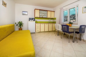 A compact room with a yellow bed, a small kitchenette, a dining area with two chairs, and a window with a view of the outdoors. - Apartment Seafront Apartments (Rogoznica)