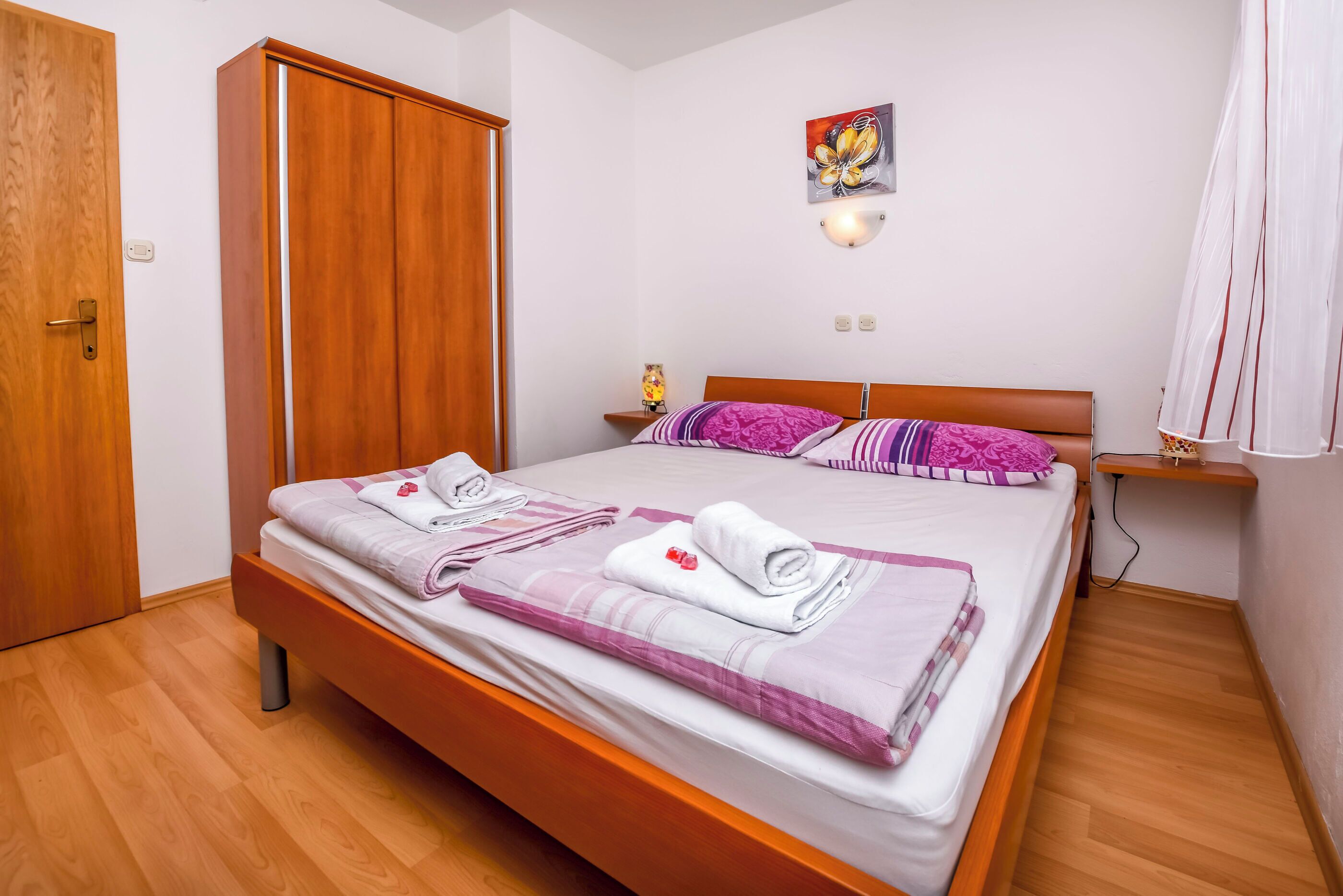 A neatly made bed with towels, a wooden wardrobe, a bedside table with a lamp, and a wall-mounted light fixture.