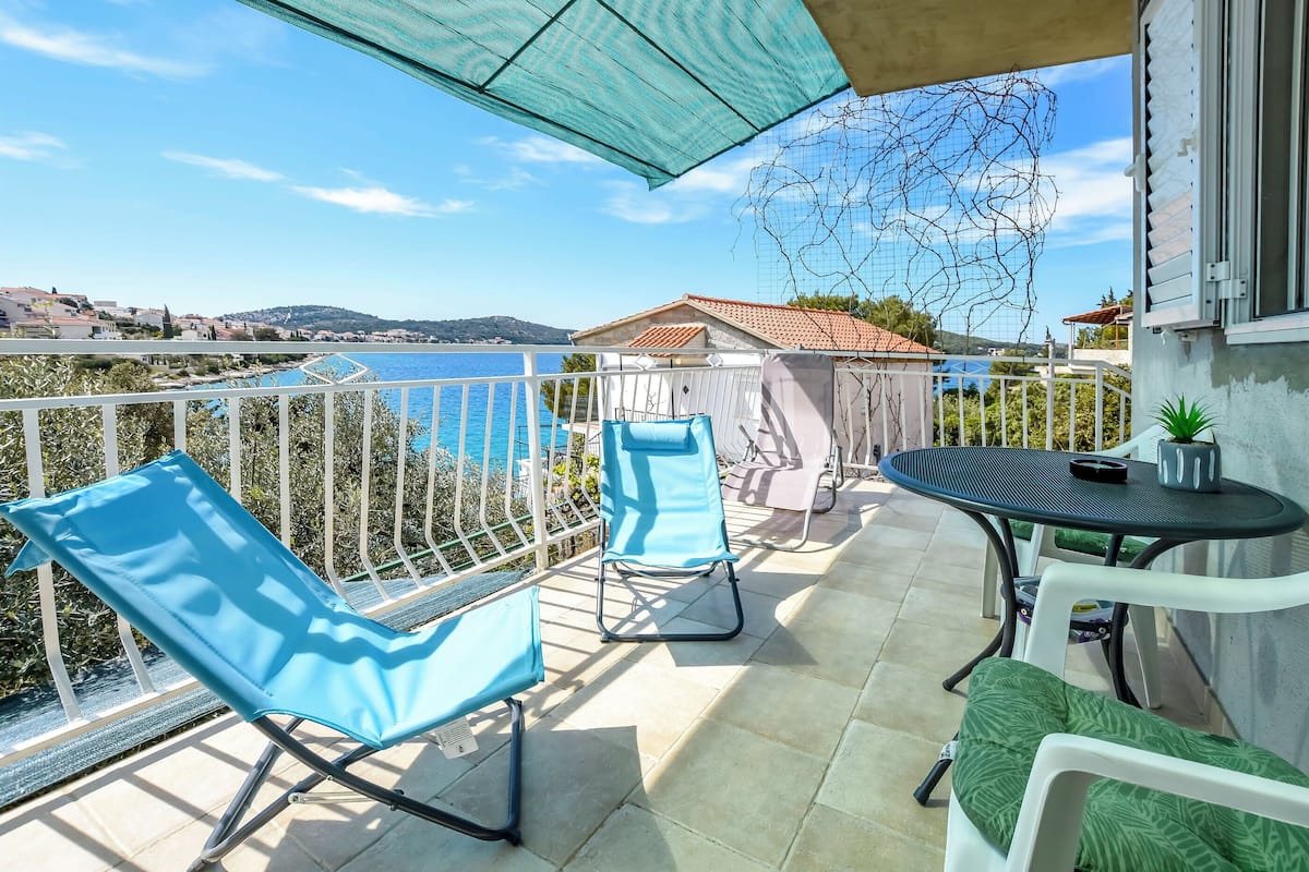 Un balcón con dos sillas azules, una mesa pequeña con una silla y vistas al mar y las colinas.