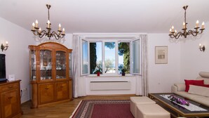 A living room with a wooden cabinet, a sofa, a coffee table, and a large window with a view of trees. - Apartment Cypress (Okrug Gornji)