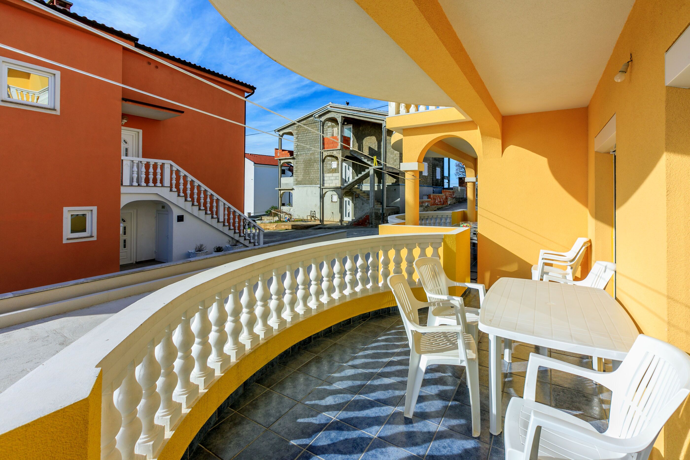 Un balcone con sedie e tavolo bianchi, una parete gialla e un edificio rosso sullo sfondo.