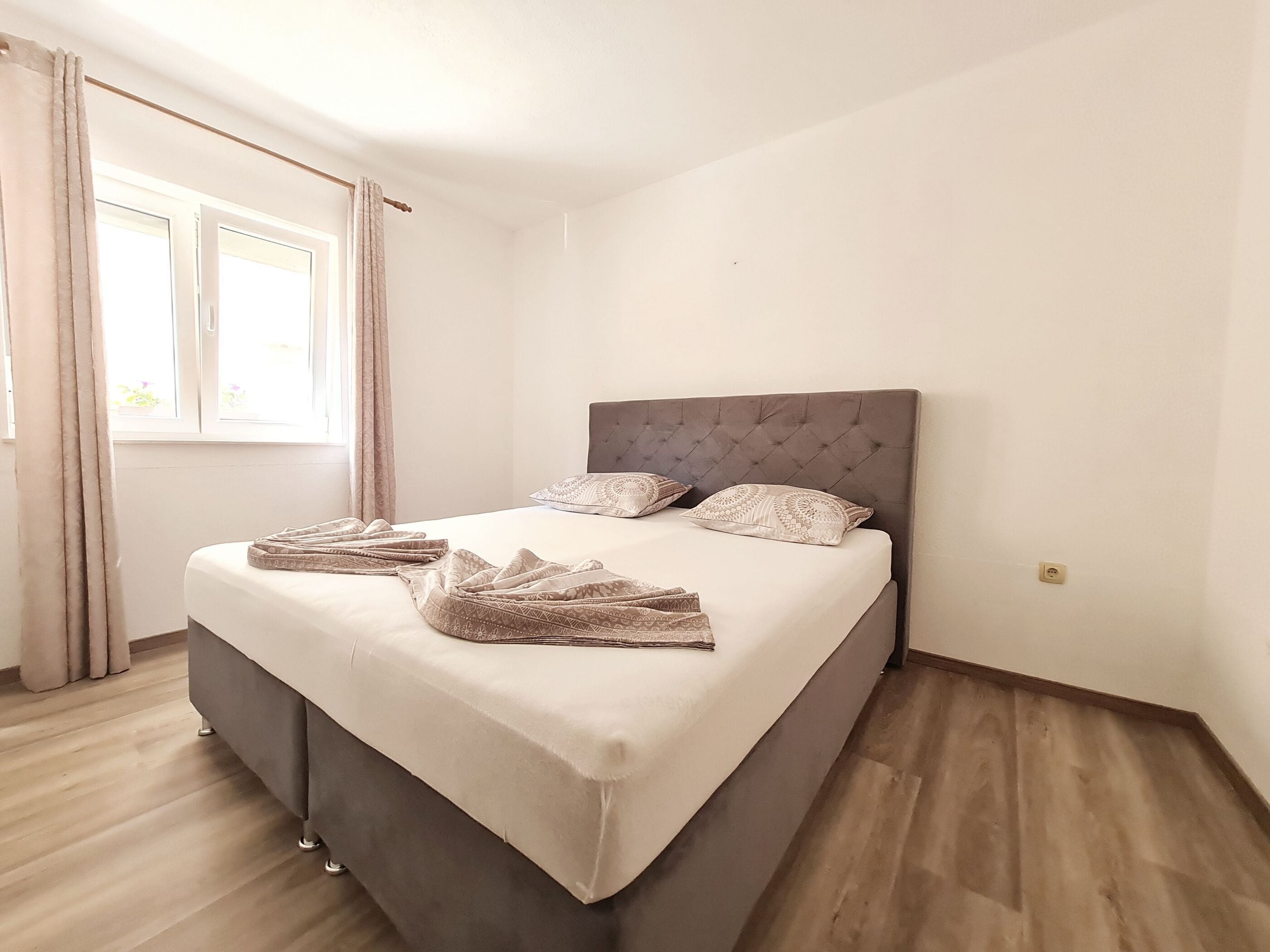 A neatly made bed with a grey headboard, a window with curtains, and a wooden floor.