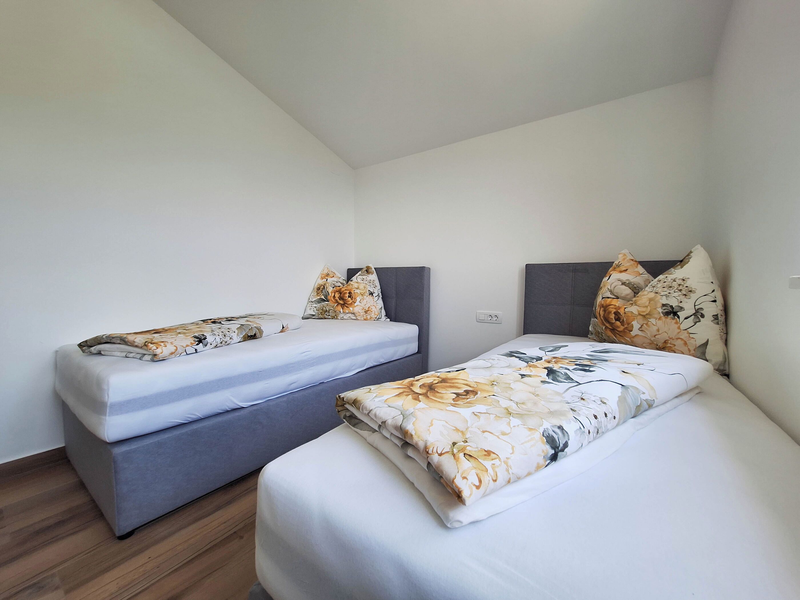 Ein Zimmer mit zwei Betten, jeweils mit blumigem Bettbezug und Kissen, grauen Kopfteilen und einem Holzboden.
