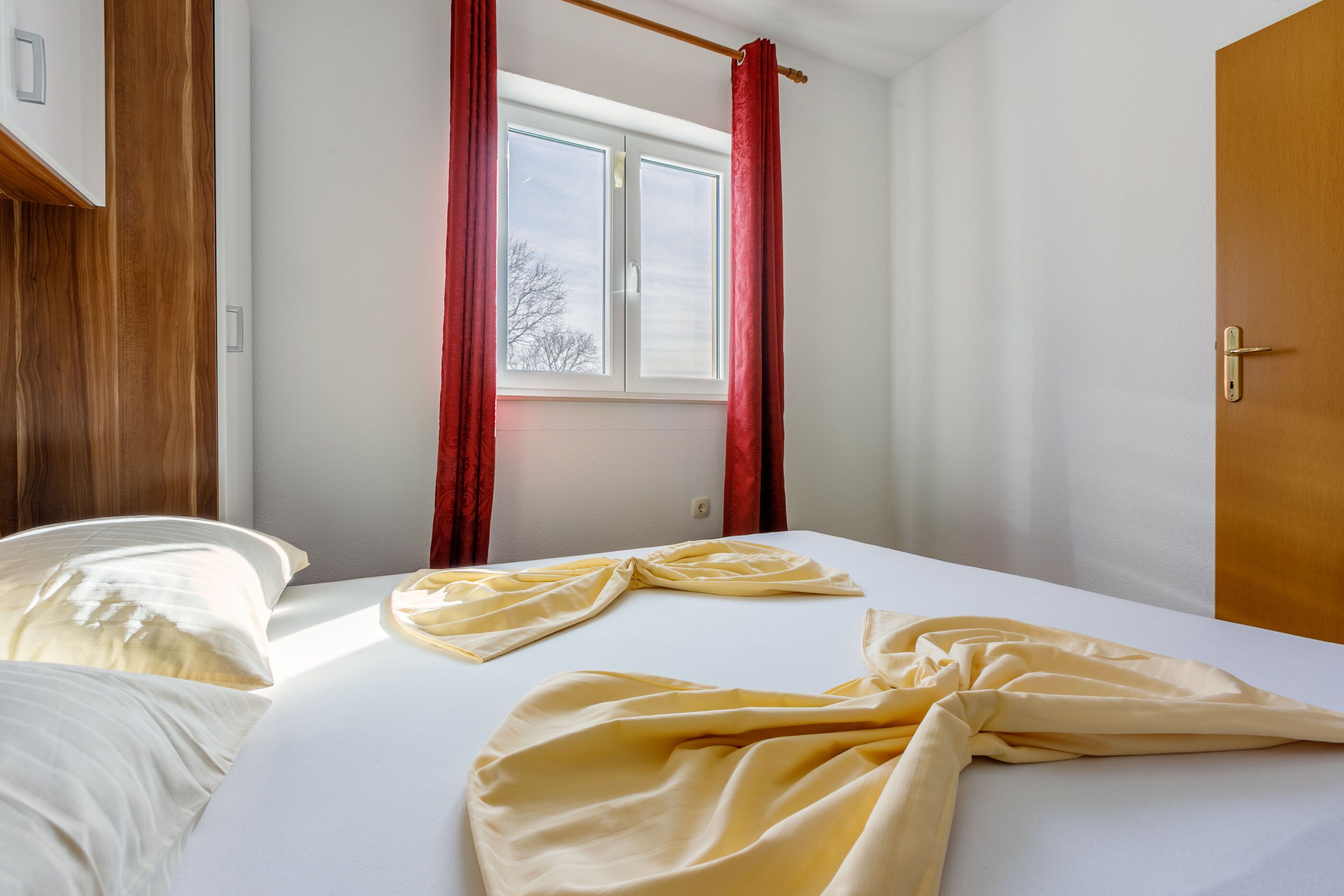 A hotel room with a bed, a window with red curtains, and a wooden door.