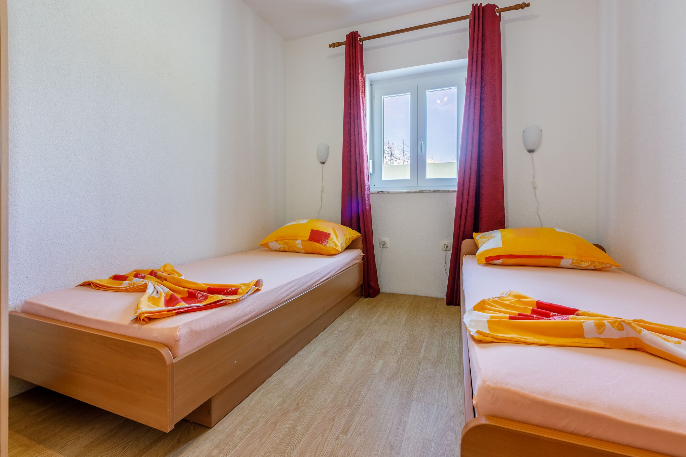 A room with two single beds, wooden flooring, a window with curtains, and a wall-mounted light fixture.