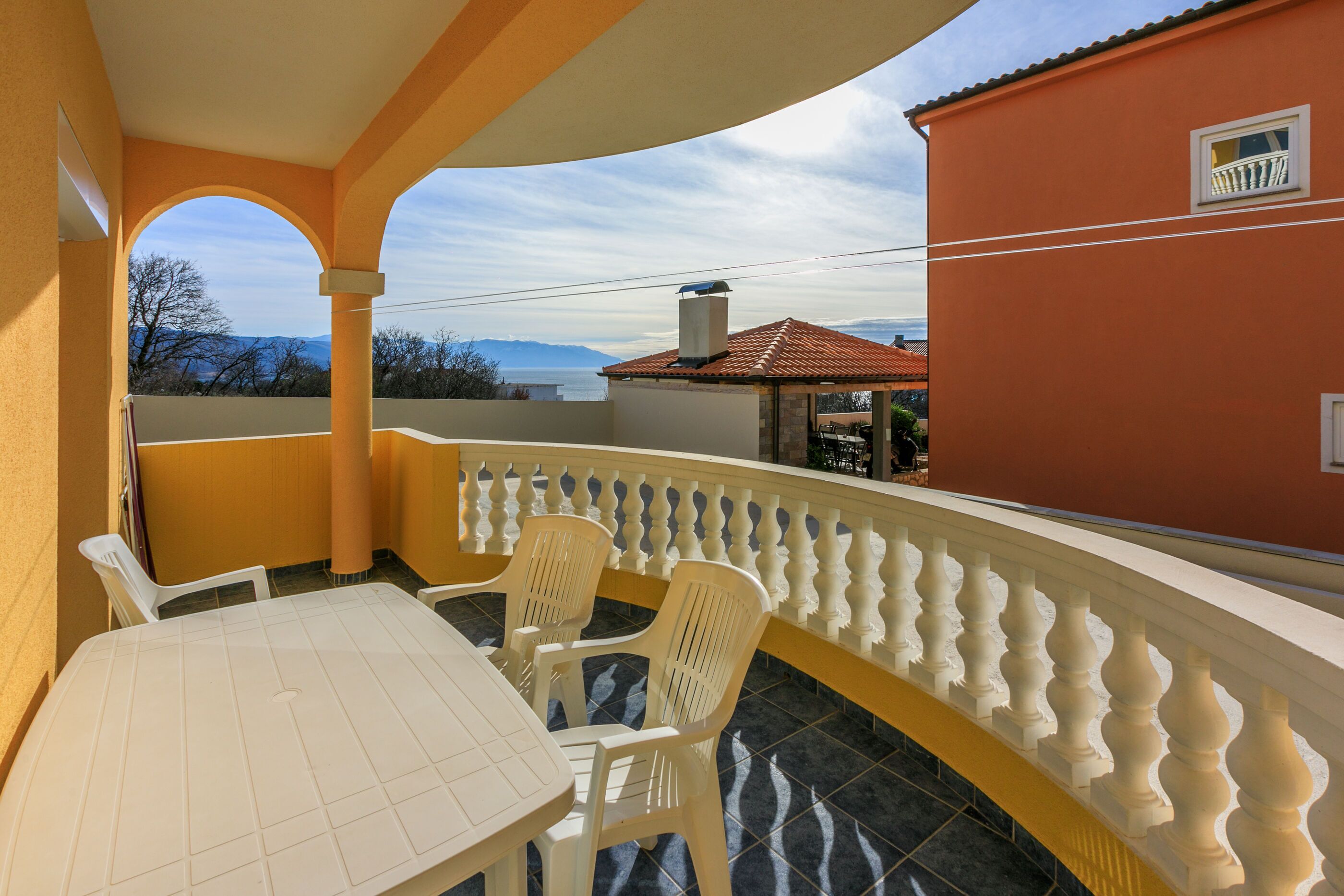 A balcony with a table and chairs, a railing, and a view of a building and mountains.