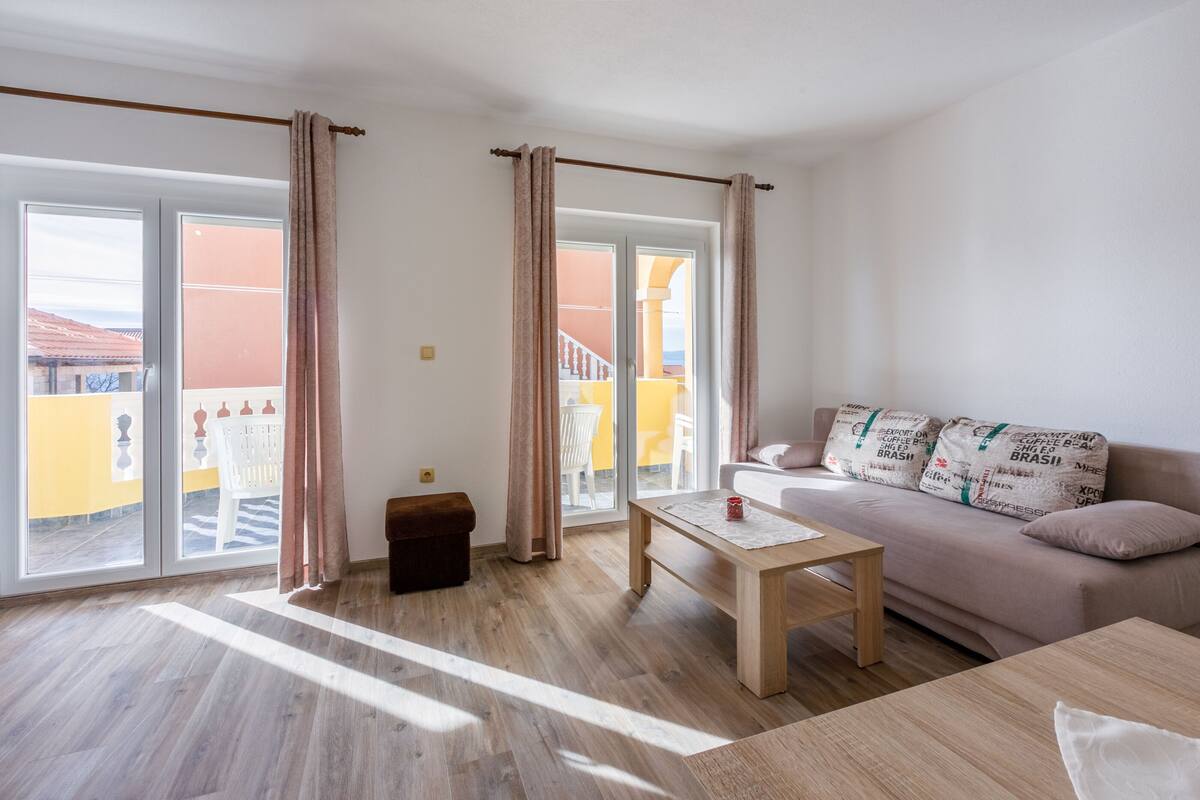 A modern living room with a sofa, a wooden coffee table, and a bench. Large windows with curtains allow natural light and a view of the outside.