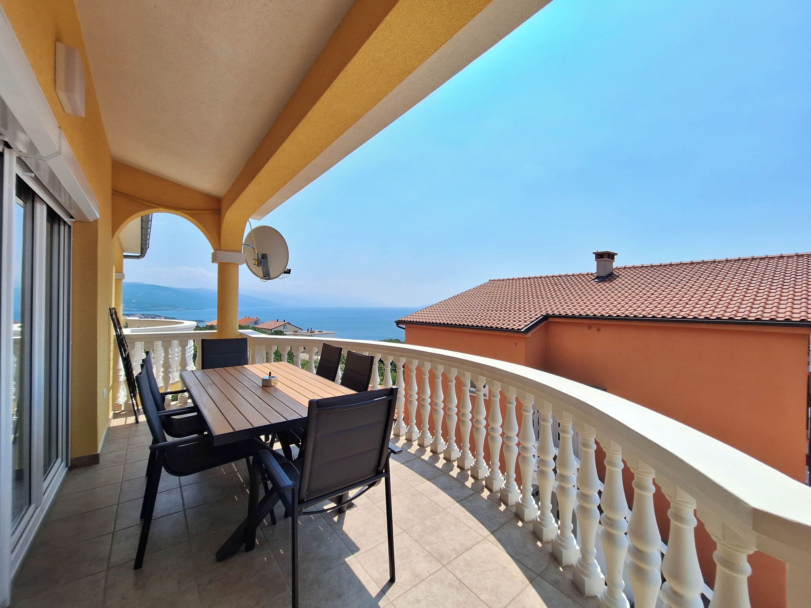 A balcony with a wooden table and chairs, a white balustrade, and a view of the sea and distant mountains.