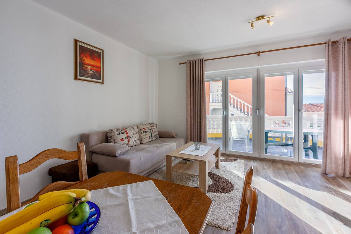 A living room with a sofa, a dining table with fruits, a coffee table, and a view of a balcony with outdoor furniture.