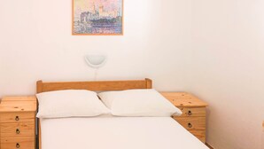 A neatly made bed with white linens and two pillows, a wooden nightstand with drawers, a small table with a white lamp, and a framed painting of a cityscape on the wall. - Apartment Donna (Biograd)