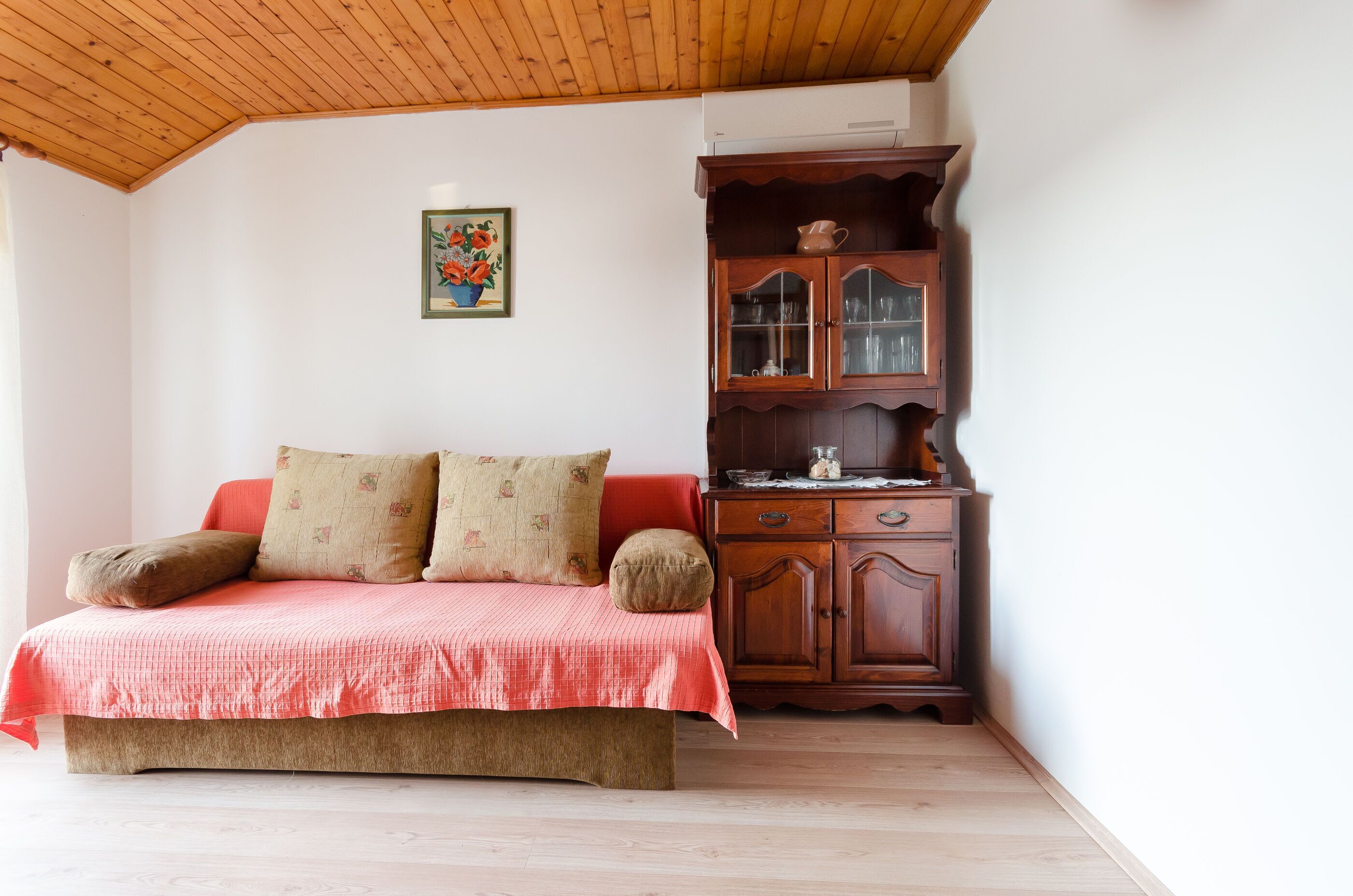A bedroom with a wooden ceiling, a bed with a red bedspread, a wooden cabinet, and a framed picture on the wall.