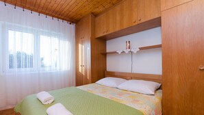Una stanza con soffitto in legno, un letto con coperta verde, cuscini bianchi e trapunta bianca. Si vede una finestra con tende trasparenti e una mensola in legno sopra il letto.