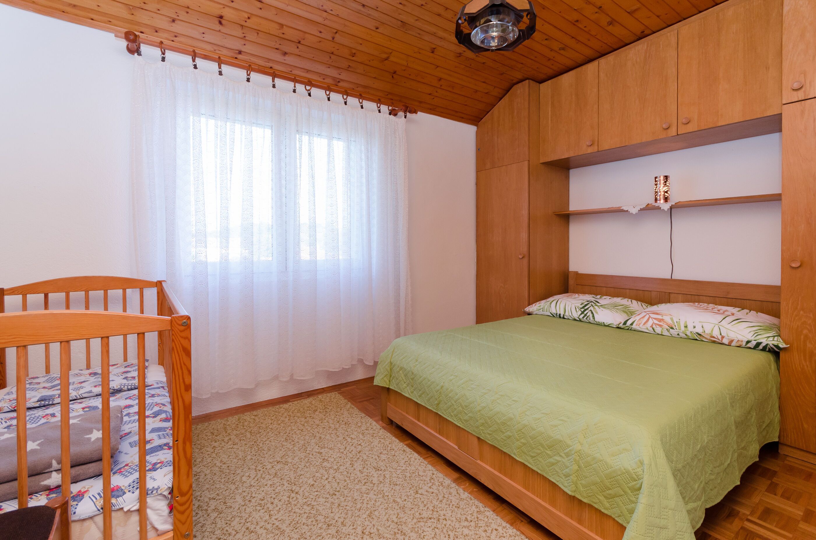 A bedroom with a wooden headboard, a bed with a green bedspread, a crib with a patterned blanket, and a window with sheer curtains.