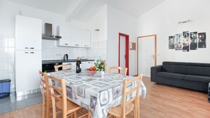 Ett modernt kök med vita skåp, ett matbord med ett mönstrat bordsduks och en grå soffa i vardagsrummet.