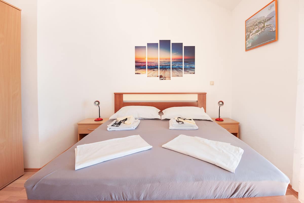 A neatly made bed with white linens, a wooden headboard, and a wall-mounted artwork depicting a sunset over the ocean.