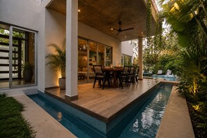A modern outdoor pool area with a dining table and chairs, surrounded by lush greenery and a clear view of the house.