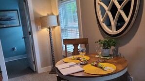 A dining area with a round table set for two, a window with blinds, and a wall-mounted decorative mirror. - Water Feature Cottage (Raleigh)