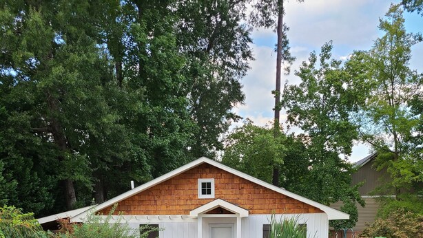 A wooden cabin with a white door and a small window, surrounded by lush greenery and a pond with lily pads. - Water Feature Cottage (Raleigh)