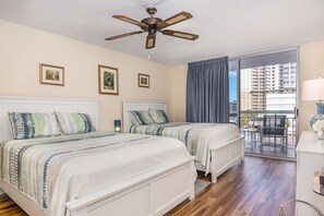 Ein Hotelzimmer mit zwei Betten, einem Deckenventilator, einem Balkon mit Blick auf Gebäude und einem Schreibtisch mit Lampe.