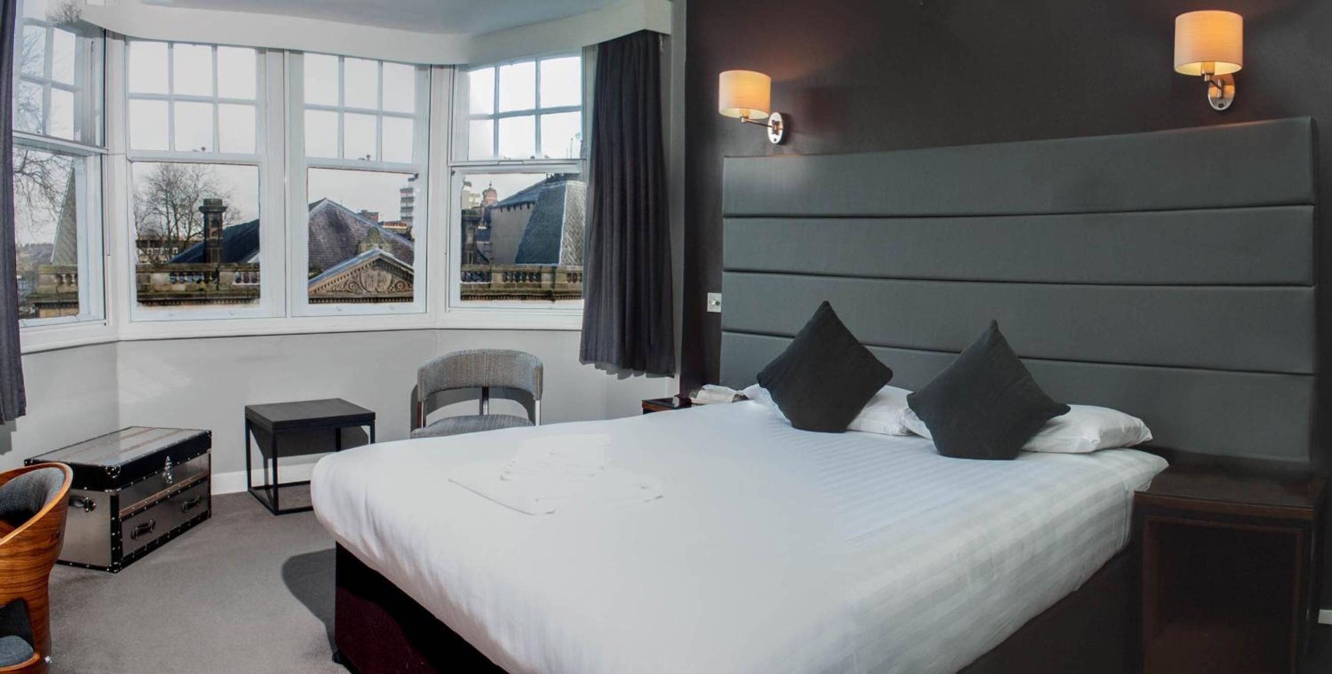 A hotel room with a large bed, two bedside lamps, a chair, a small table, and a window with a view of buildings.