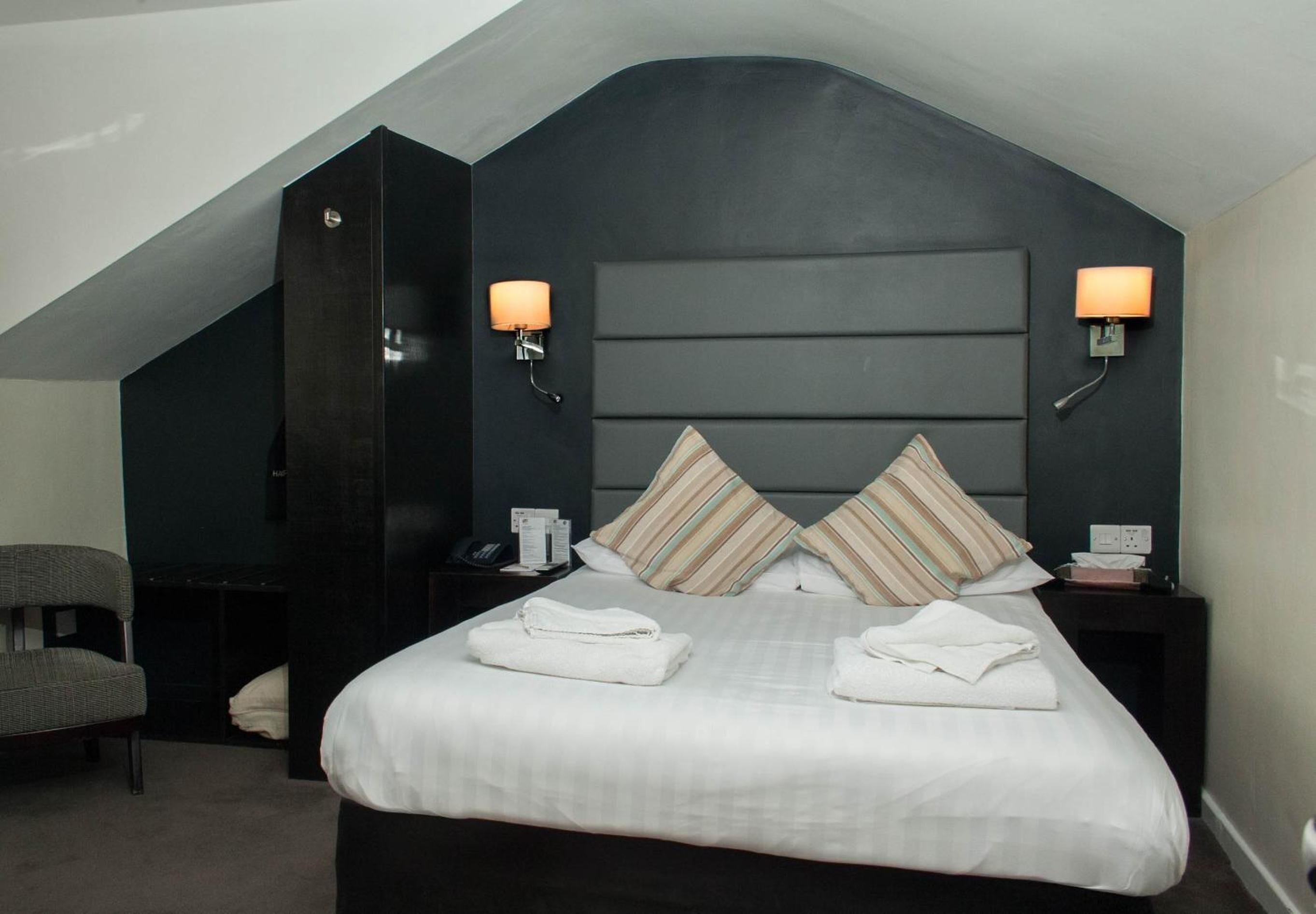 A modern hotel room with a large bed, two bedside tables, a chair, and a dark accent wall.