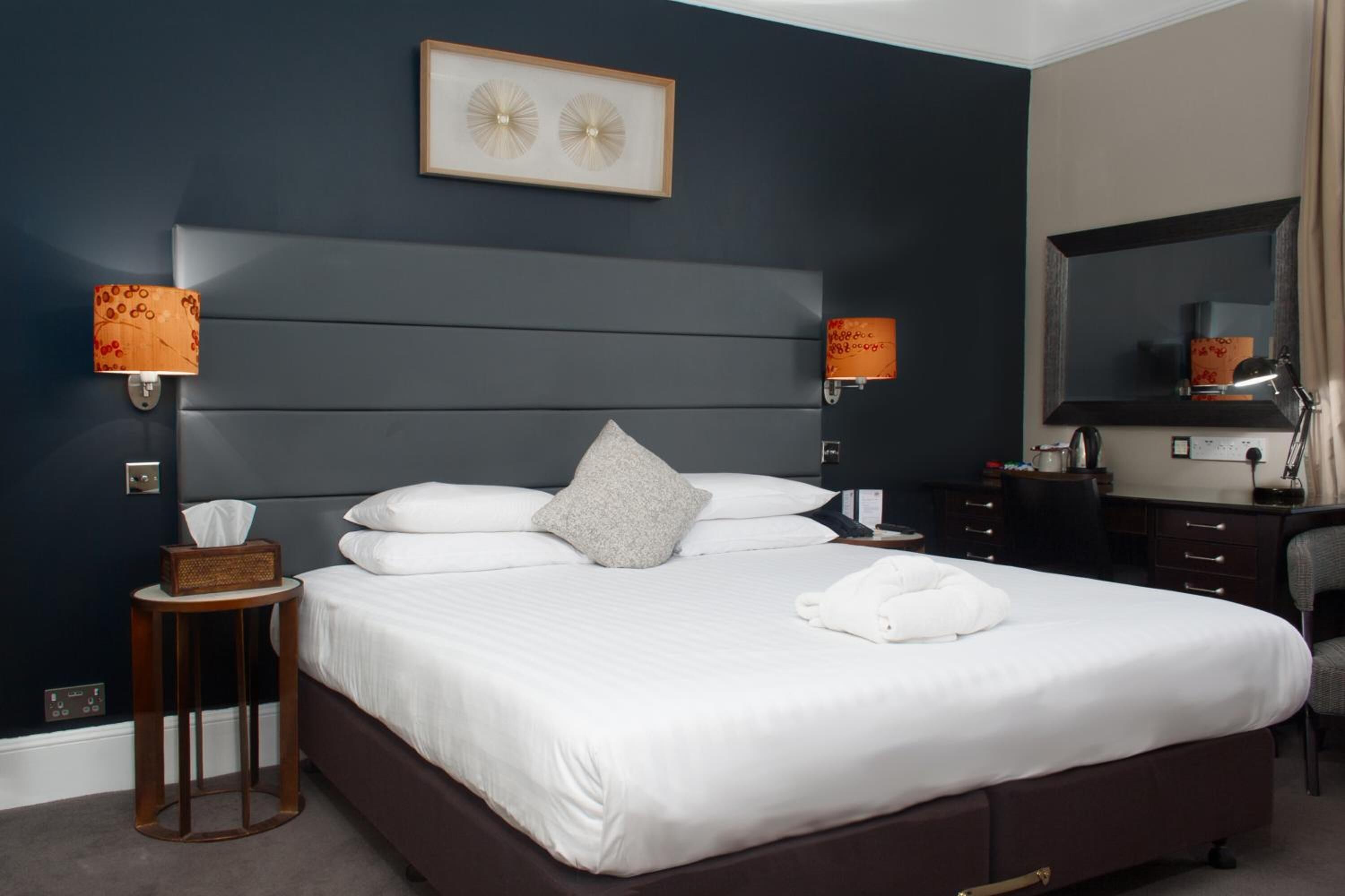 A neatly made bed with white linens, a grey pillow, and a white towel on the side. There are two wall-mounted lamps with orange shades on either side of the bed. A framed picture hangs above the bed, and a mirror is on the wall to the right.