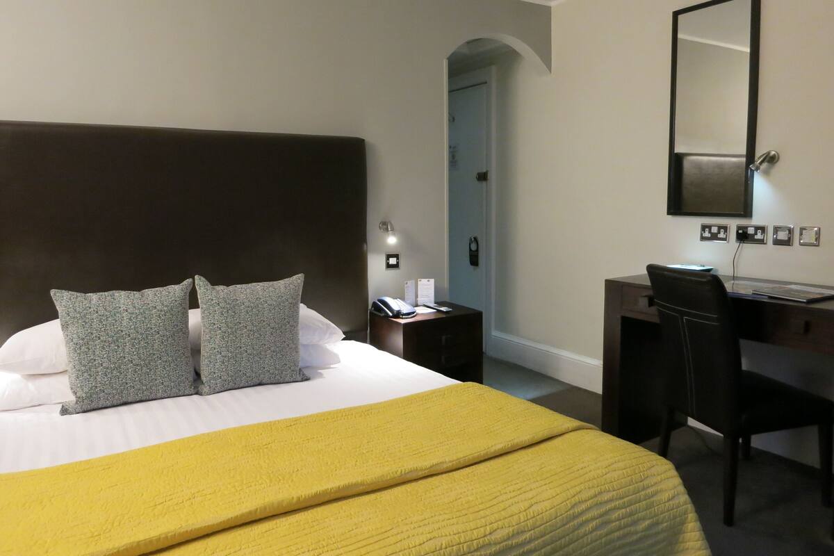 A hotel room with a large bed, two pillows, a desk with a chair, a telephone, and a mirror on the wall.