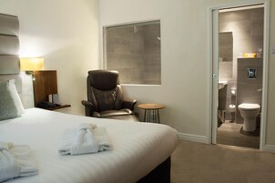 A hotel room with a large bed, a leather chair, a small round table, and a bathroom visible through an open door.