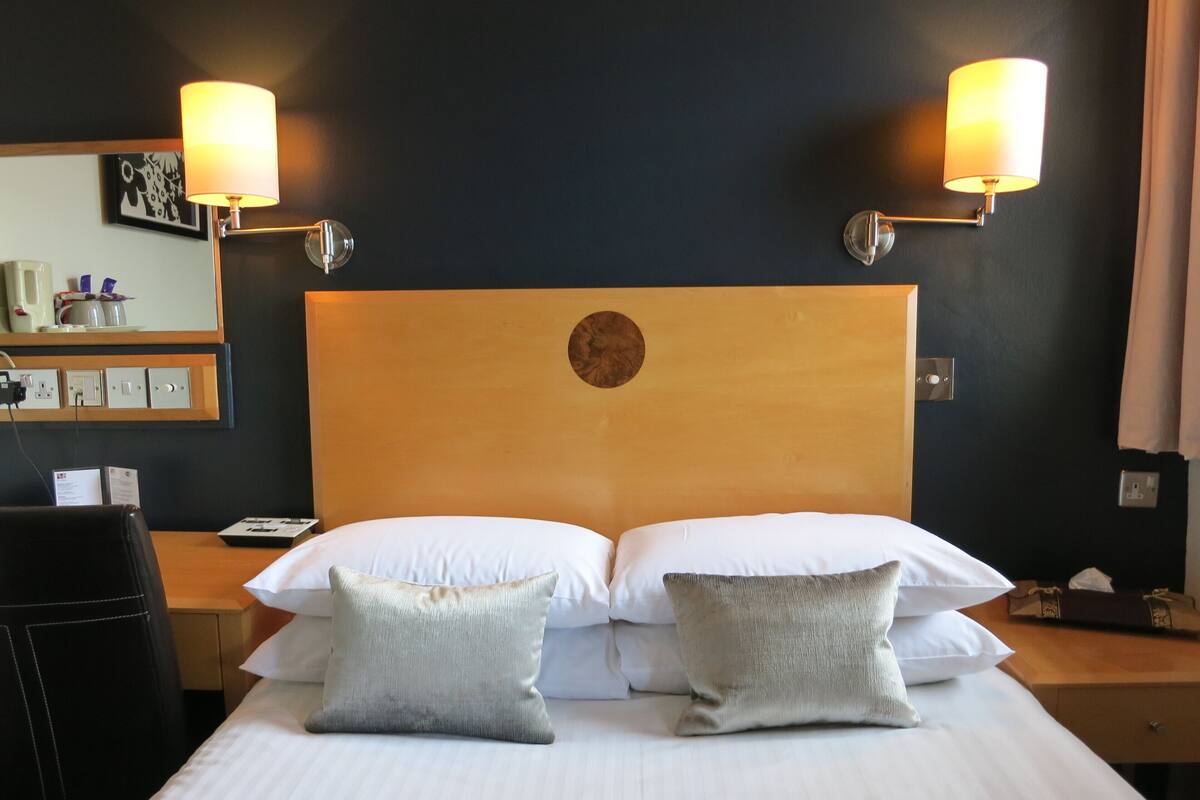 A hotel room with a large bed, two bedside lamps, a wooden headboard, and a small table with a lamp.