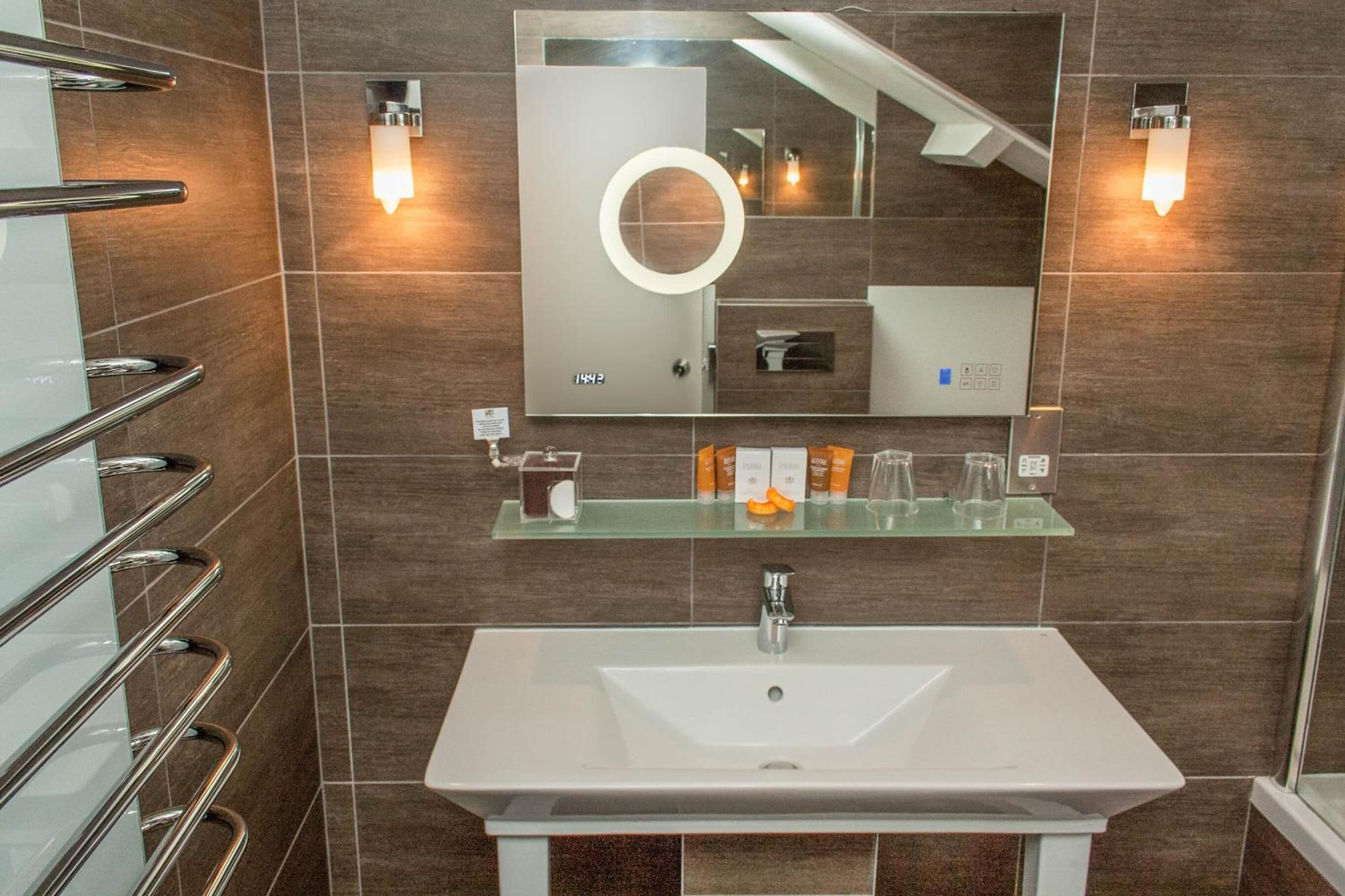 Un baño moderno con un lavamanos blanco, repisas de vidrio, un espejo grande con luz circular y dos lámparas de pared.