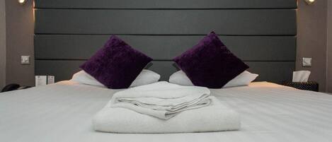 A neatly made bed with white linens and two purple pillows, a dark headboard, and bedside lamps.