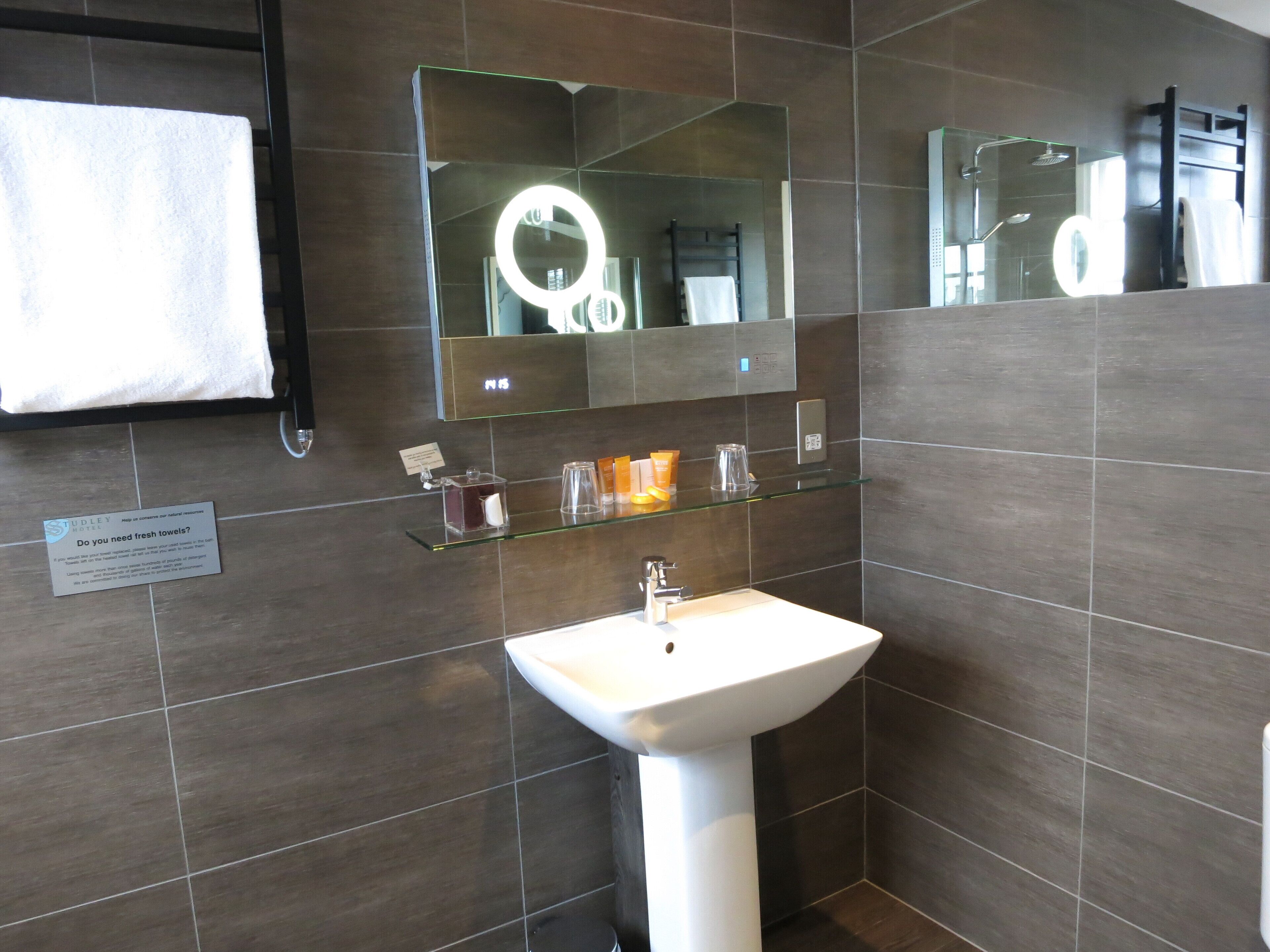 A modern bathroom with a white pedestal sink, a glass shelf with toiletries, a large mirror with a light, and a towel rack.