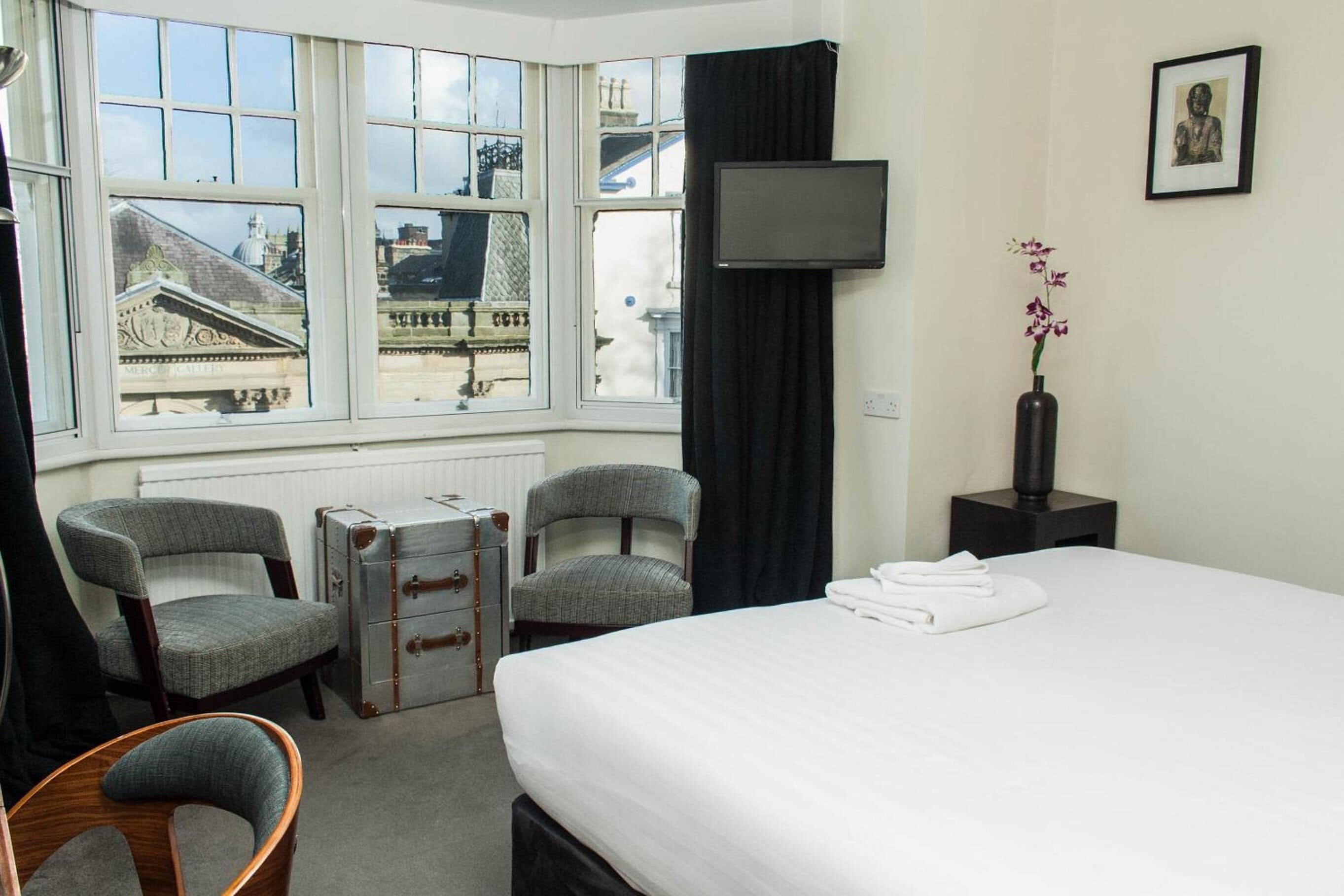 A hotel room with a bed, two armchairs, a desk, a television, and a window with a view of buildings.