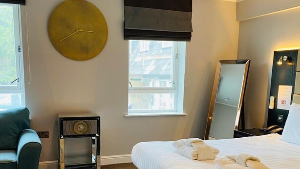 Habitación de hotel con cama, una silla, una mesita, un reloj, una ventana y un espejo.